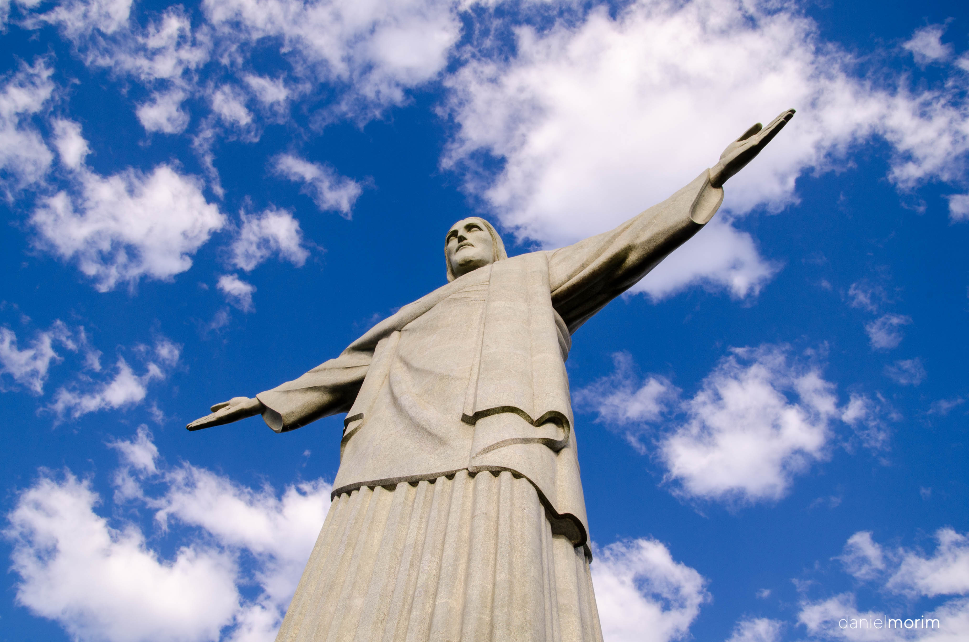 Cristo Redentor visto de baixo, com os bra&ccedil;os abertos e o c&eacute;u azul ao fundo